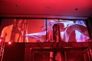 a black dj with long braids stands at their deck with harsh gray and red visuals projected behind them