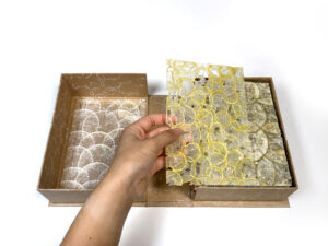 a left-hand picks up a delicate piece of paper from a box. The page is made form sliced cylindrical yellow-skinned fruit. More paper in the box ranges in color from white to green.