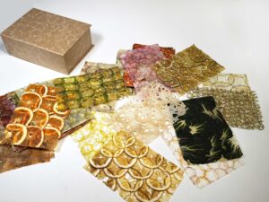 samples of paper made from thin slices of circular fruit and vegetables are piled on a white surface. The fruit is orange, yellow, green, fuchsia, and dark green.