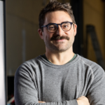 masculine person with brown mustache hair and glasses arms crossed in a grey long sleeve shirt