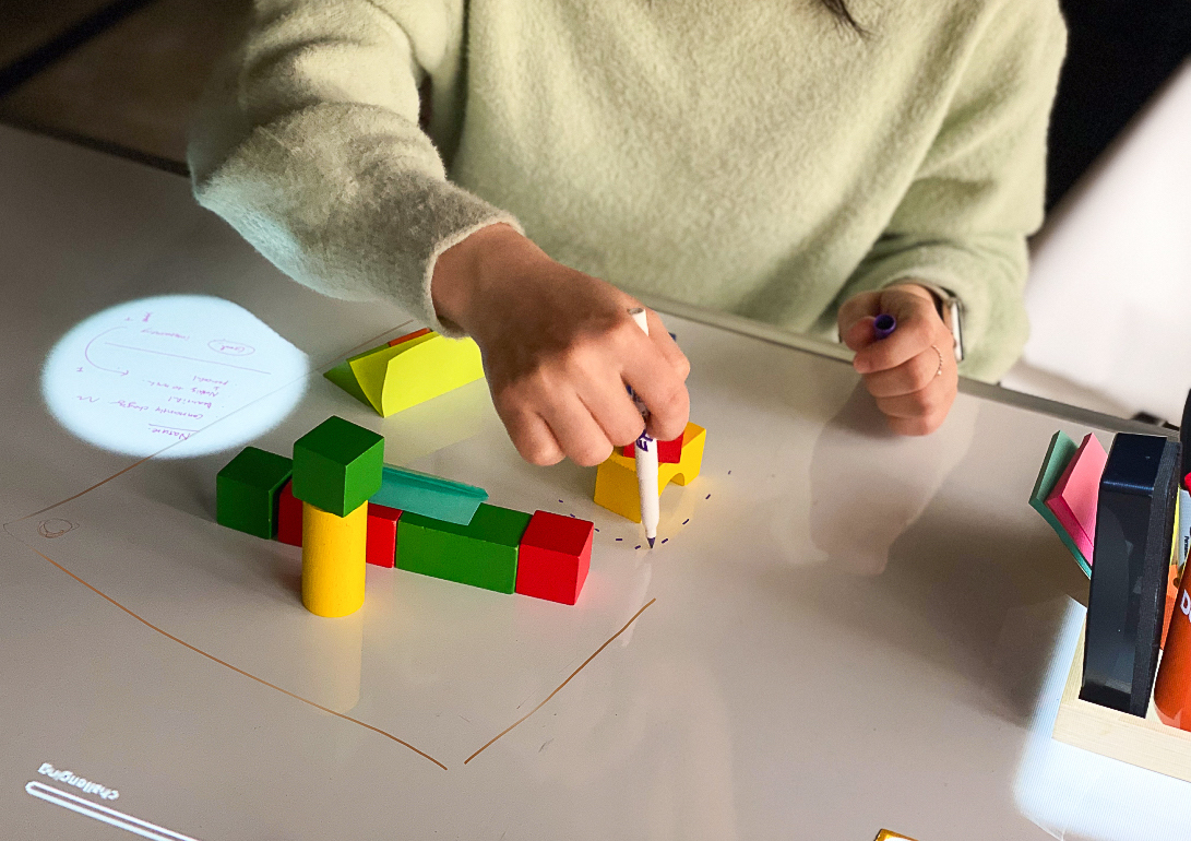 a person is seen from shoulders to waist sitting at a whiteboard table, drawing with a white board marker around wooden blocks, an interactive projection highlights writing on the board to the side of the blocks.